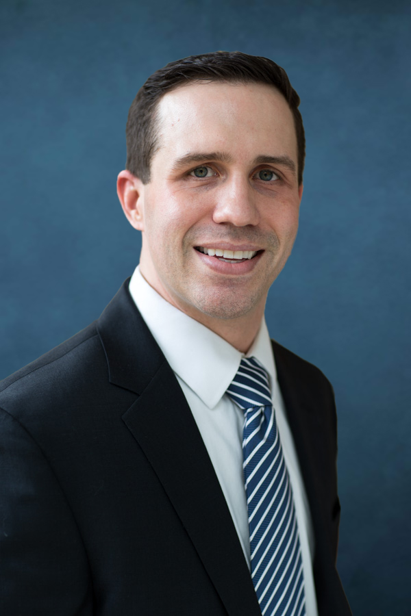 Professional portrait of Mark Pulver, PA-C, MPAS – Millcreek Dermatology Mark Pulver, PA-C, MPAS, from Millcreek Dermatology, smiling in a professional headshot while wearing a dark suit and striped tie against a blue background – Pulver