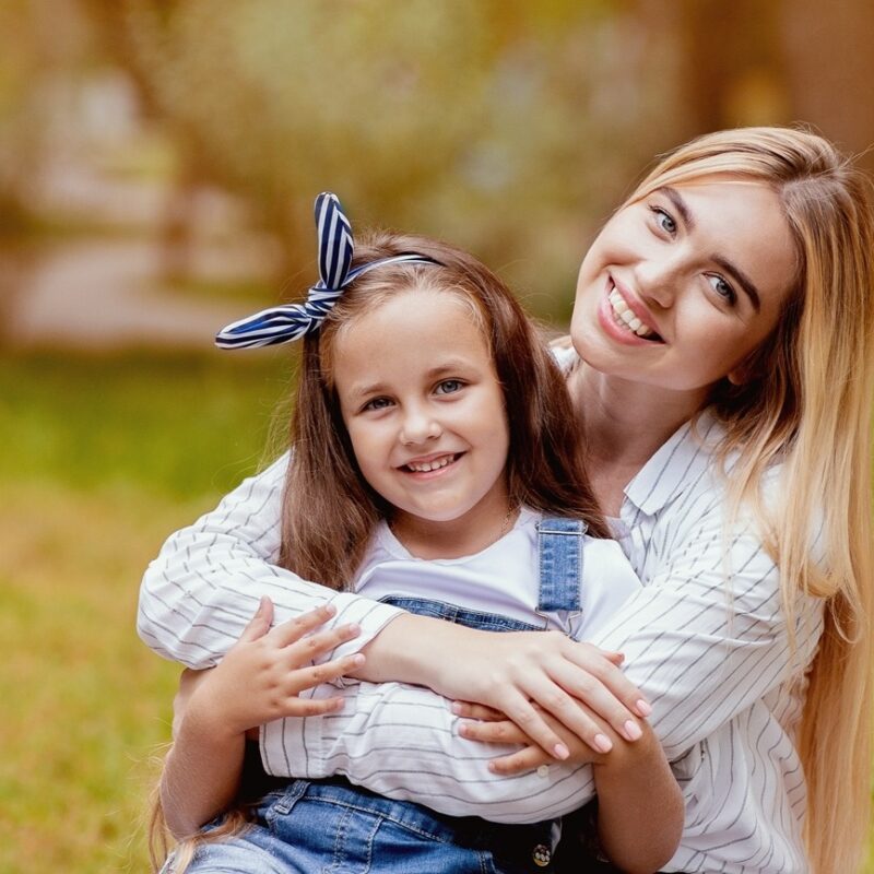 A smiling mother embraces her young daughter while sitting together on the grass in a sunlit park, representing the importance of family health and awareness of the early sign of skin cancer for prevention and protection – early sign of skin cancer
