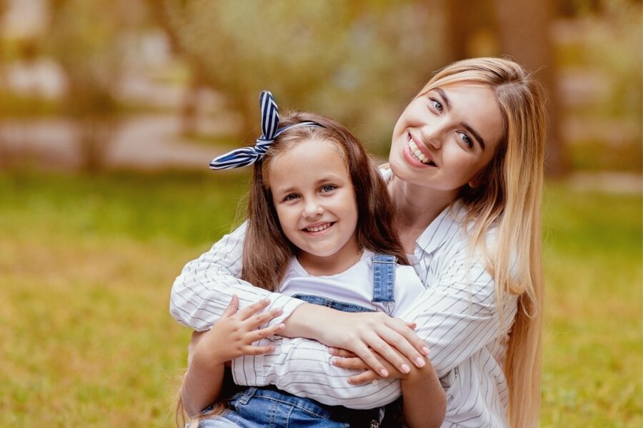 A smiling mother embraces her young daughter while sitting together on the grass in a sunlit park, representing the importance of family health and awareness of the early sign of skin cancer for prevention and protection – early sign of skin cancer