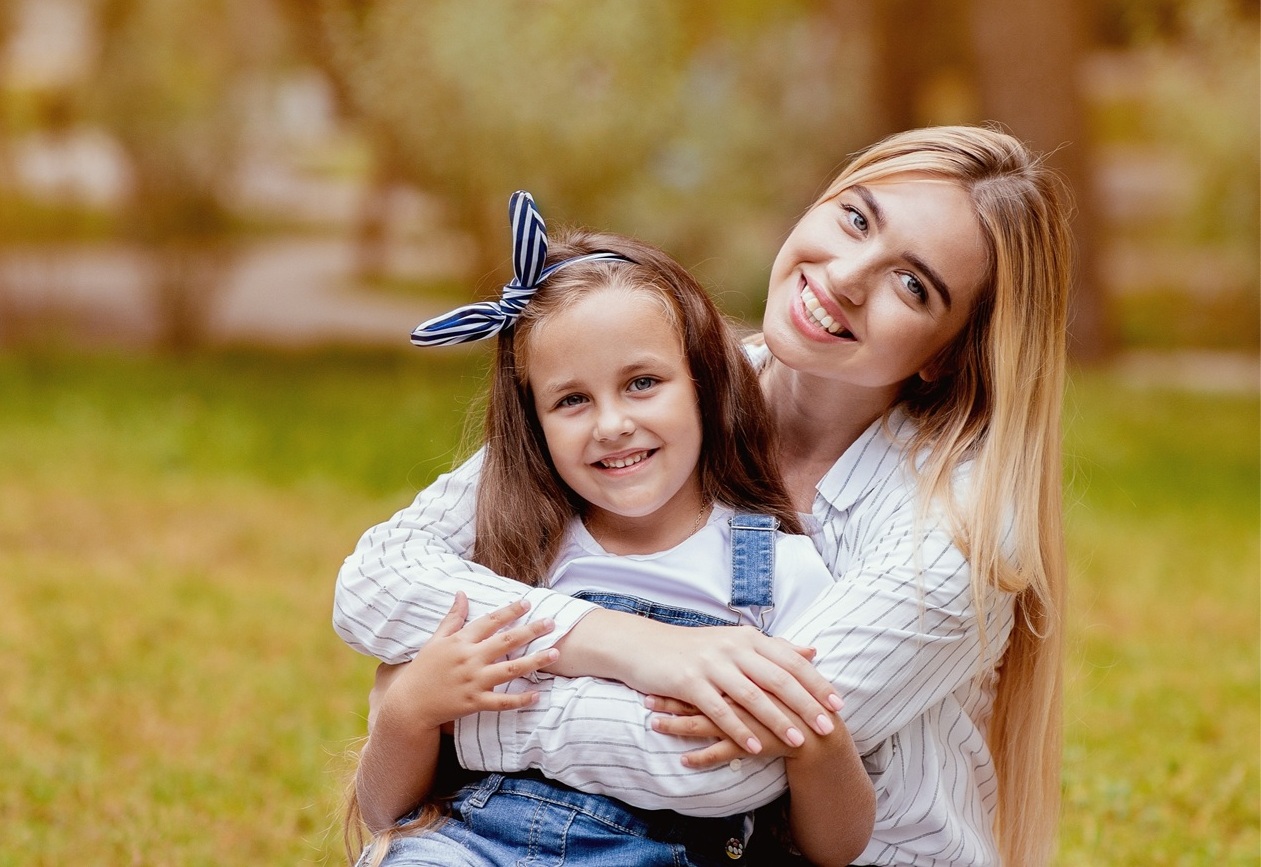 A smiling mother embraces her young daughter while sitting together on the grass in a sunlit park, representing the importance of family health and awareness of the early sign of skin cancer for prevention and protection – early sign of skin cancer