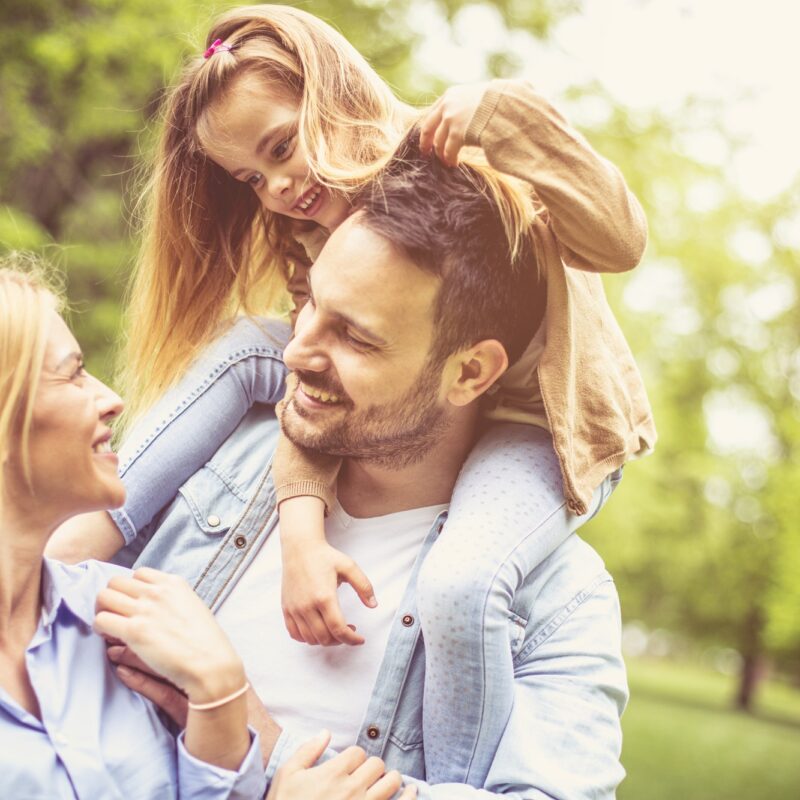 Smiling man carries a young girl on his shoulders while standing with a woman in a sunlit park, symbolizing healthy, confident living and the role dermatologists play in supporting skin health for the whole family – what does a dermatologist do