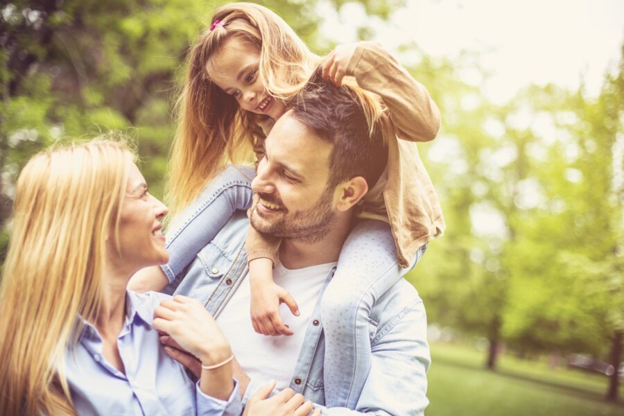 Smiling man carries a young girl on his shoulders while standing with a woman in a sunlit park, symbolizing healthy, confident living and the role dermatologists play in supporting skin health for the whole family – what does a dermatologist do
