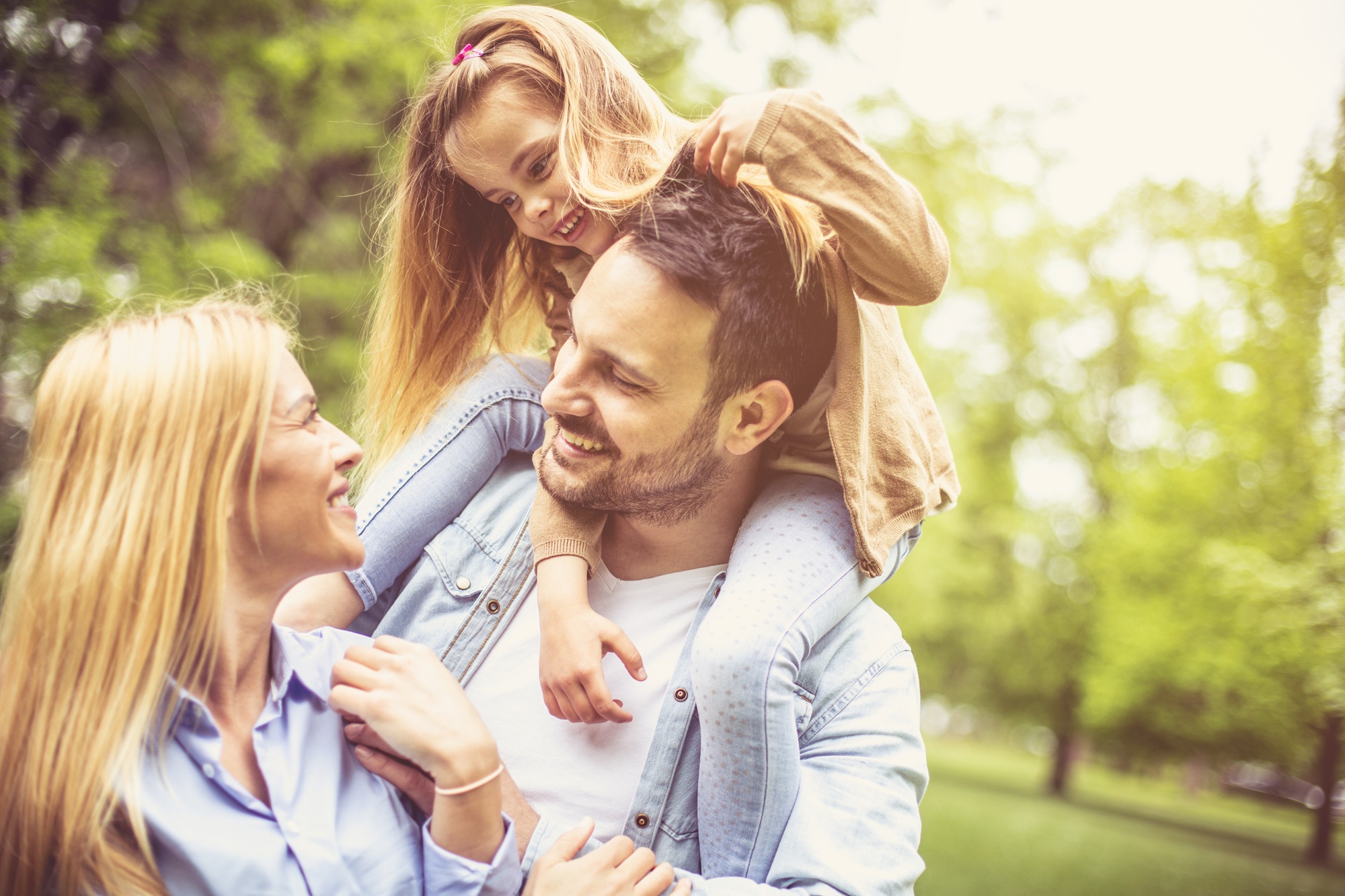 Smiling man carries a young girl on his shoulders while standing with a woman in a sunlit park, symbolizing healthy, confident living and the role dermatologists play in supporting skin health for the whole family – what does a dermatologist do