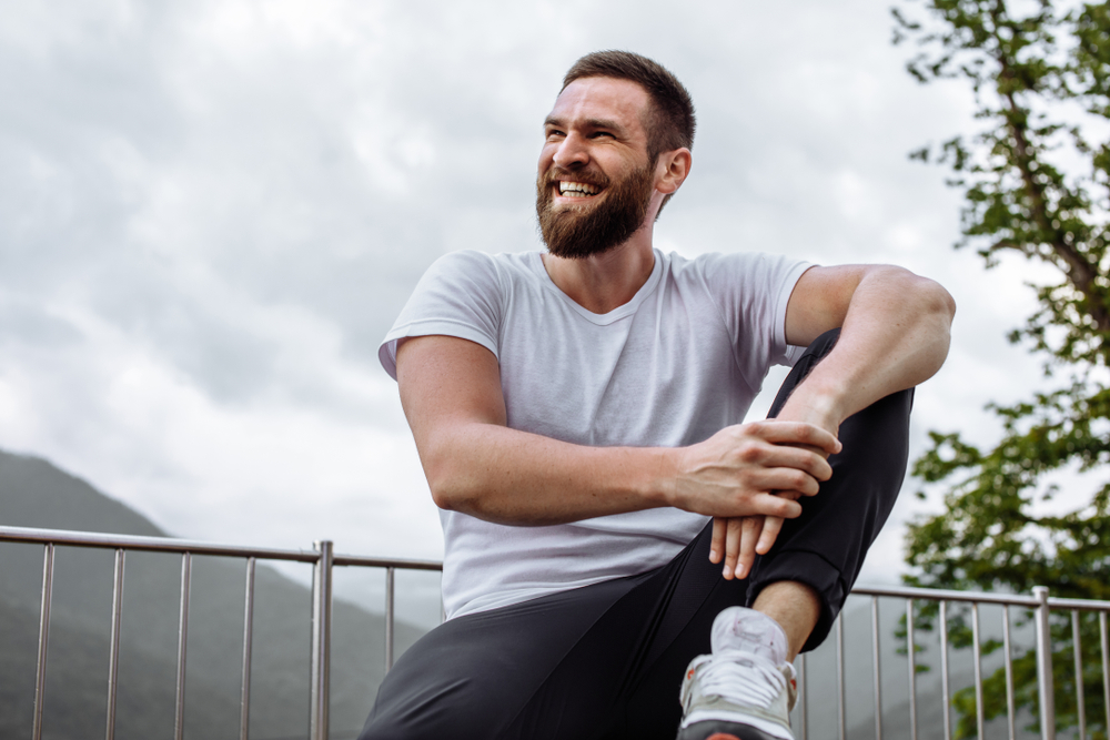 Bearded man in a white t-shirt and black athletic pants sits relaxed outdoors on a railing with mountains in the background, smiling confidently and radiating health and well-being – Millcreek Dermatology